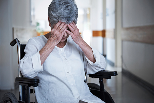 Shot of a senior woman in a wheelchair sitting with her hands over her eyes in the hallway of a nursing home.