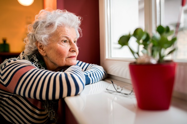 An elderly woman with white hair wearing a striped shirt rests her arms on a windowsill inside a Georgia nursing home, staring outside thoughtfully.