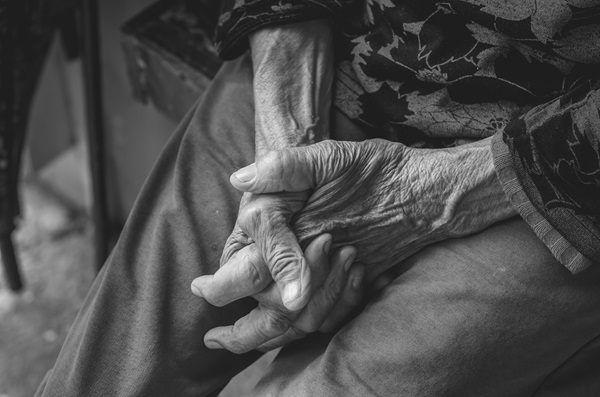 A black-and-white, close-up shot of an elderly person's thin, weathered hands clasped tightly together in their lap.
