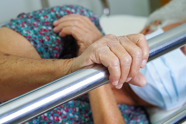A close-up of an elderly patient’s hand, wearing a blue floral gown and a surgical mask, holding onto the metal side rail of a bed in a nursing home.